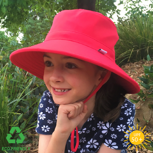 A child wearing a watermelon coloured Eco Friendly bucket hat with a wide brim, sitting outdoors with a garden and sunlight in the background.